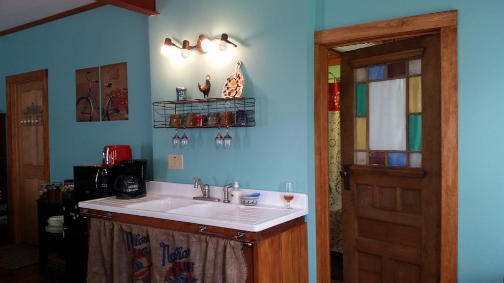 A kitchenette features a wooden countertop with a sink and a variety of kitchen tools. Colorful dishware is displayed on a wall-mounted rack. The walls are painted in a soft teal, which complements the warm wood tones of the cabinets and door.