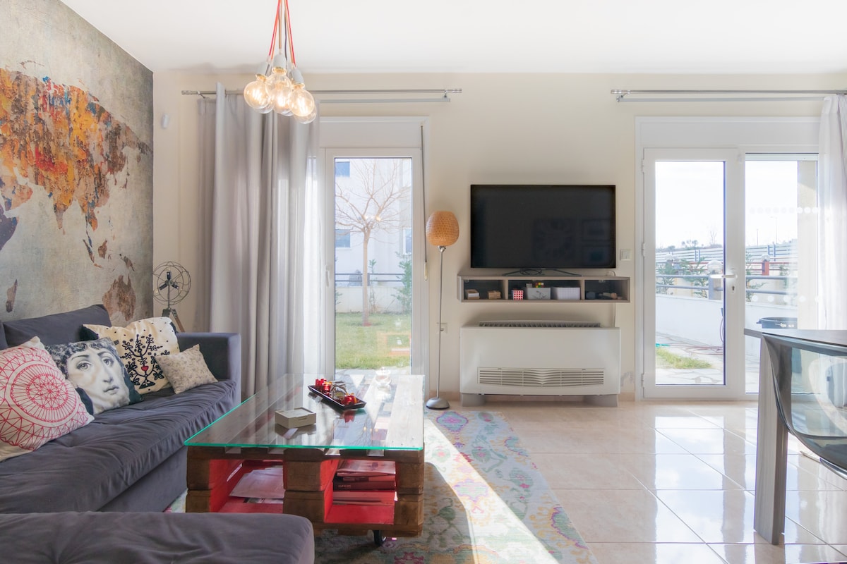 A living area is seen with a comfortable sofa adorned with patterned cushions. A low glass table sits at the center, with decorative items displayed. Large windows allow natural light to fill the space, and a flat-screen television is mounted on the wall.