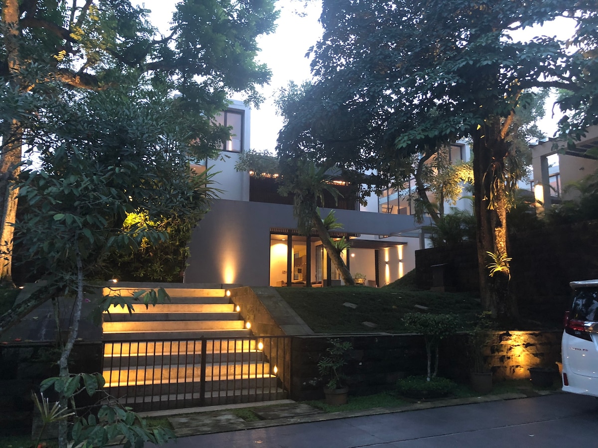 A modern architectural design is highlighted by soft lighting in the evening. Steps lead up to an entrance surrounded by lush greenery and trees, creating a serene atmosphere. The driveway is visible, alongside a parked vehicle, providing a sense of accessibility.