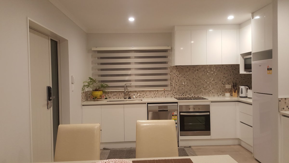 The kitchen features modern cabinetry with a clean, white finish and a textured backsplash. Equipped with stainless steel appliances, including an oven and refrigerator, it offers ample counter space. Soft lighting highlights the inviting atmosphere, while a small window allows natural light to enter.