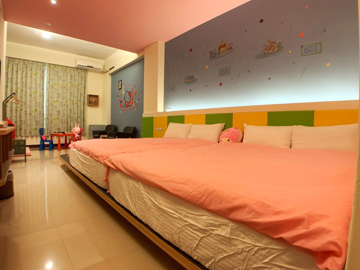 A spacious sleeping area is presented with a large bed covered by a pink blanket. Colorful wall art decorates the space, complemented by a bright ceiling. A window with light-filtering curtains and children's toys are visible, along with two chairs positioned in the corner.