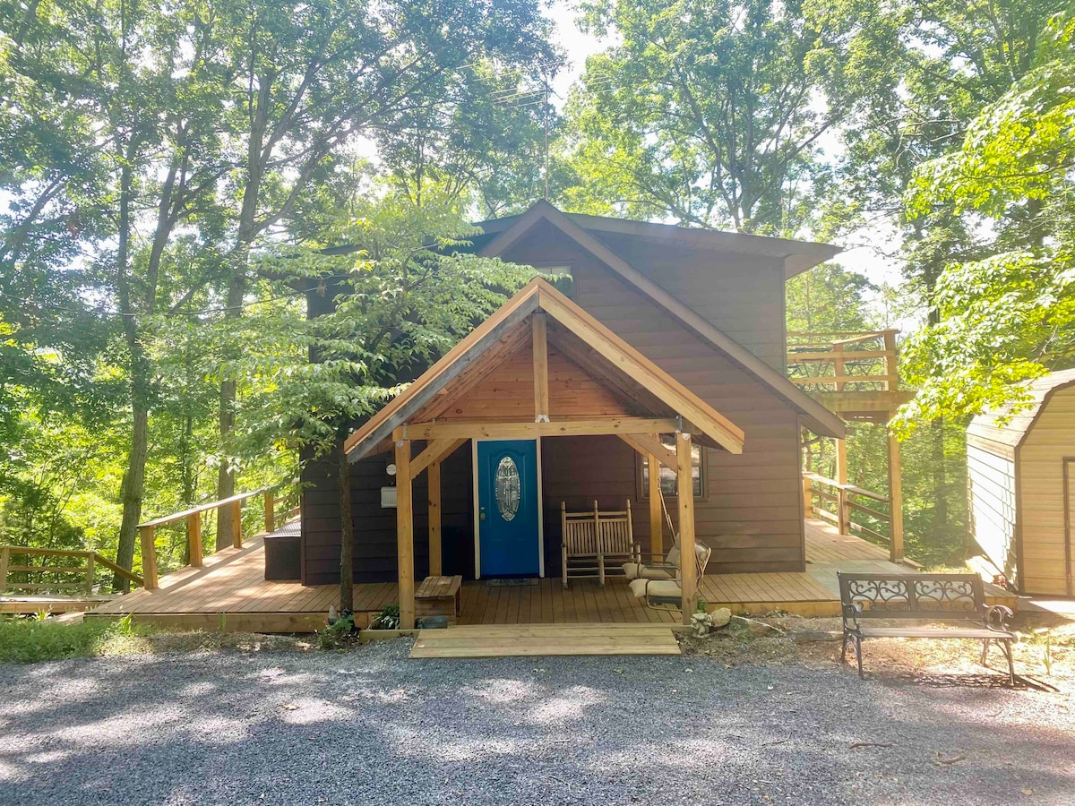 The chalet's exterior is framed by lush trees, featuring a welcoming entrance with a covered porch and rocking chairs. A blue front door adds a pop of color, while a gravel driveway leads to the entrance, ensuring easy access to the outdoor space.