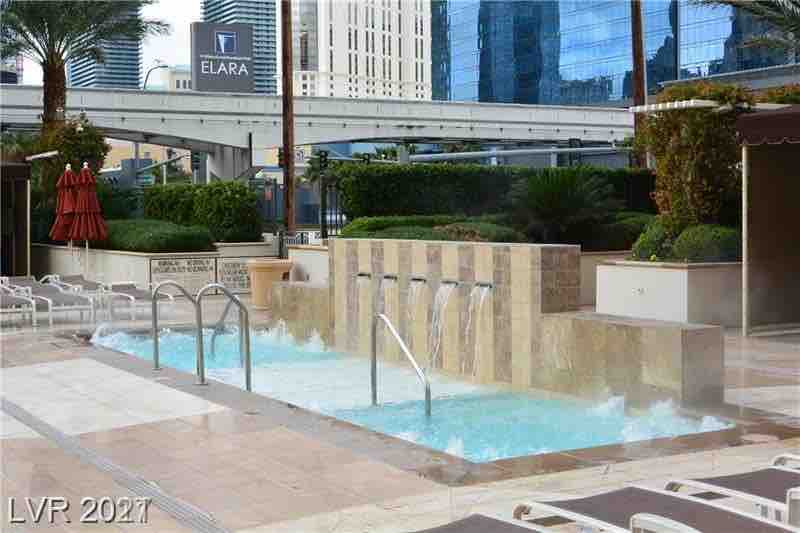 A soothing pool area features a series of vertical fountains, creating a tranquil atmosphere. The pool is surrounded by lush greenery and comfortable lounge chairs. Red umbrellas provide shade nearby, while modern high-rise buildings can be seen in the background.