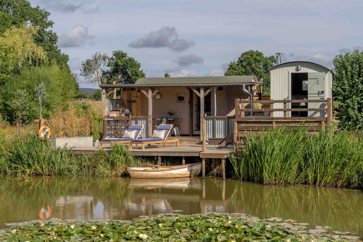 A wooden platform extends over peaceful waters, featuring a pair of lounge chairs for relaxation. An antique workman’s wagon is visible to the right. Lush green grasses border the water, enhancing the natural surroundings. A rowing boat rests quietly near the shore.