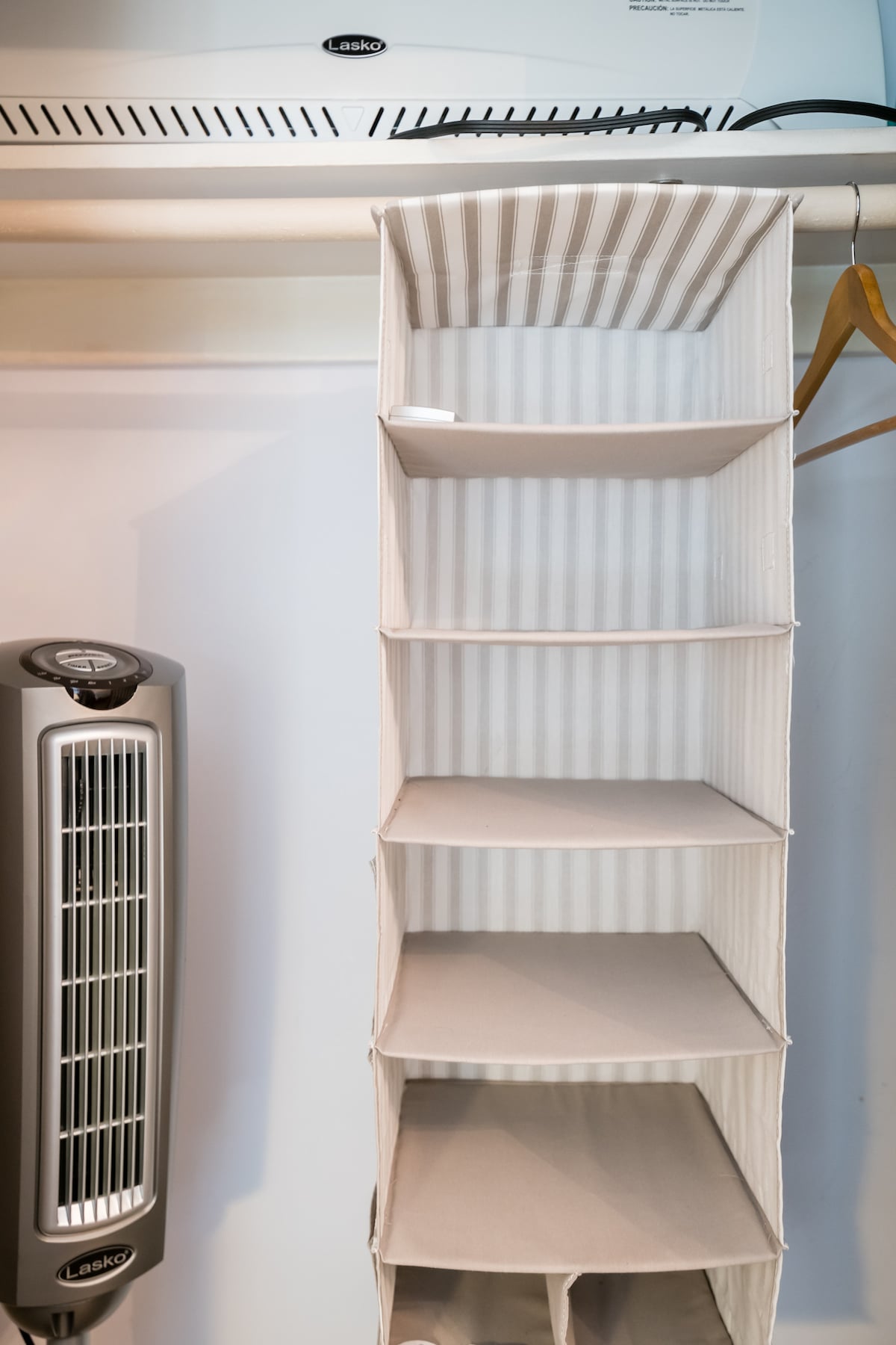 A fabric hanging organizer with several compartments is hung on a closet rod. A fan stands on the left, providing airflow, while a heater is positioned above. The light color and vertical stripes of the organizer create a tidy appearance.