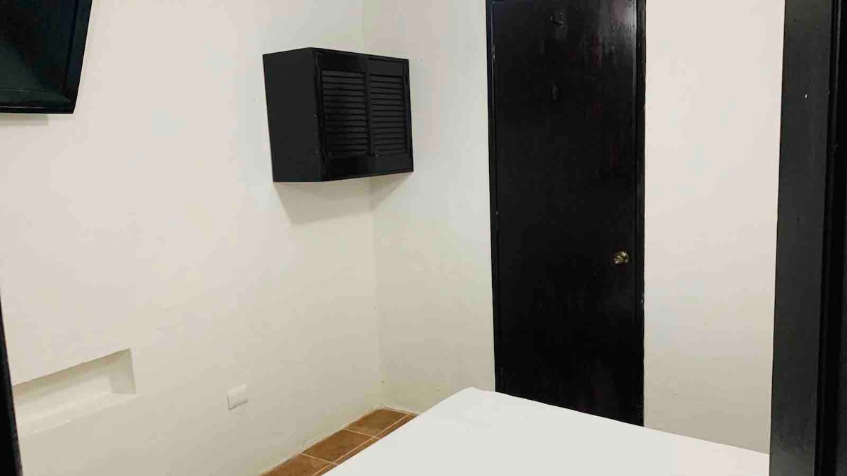 A simple bedroom space is presented, featuring a white bed and a wall-mounted television. A dark wooden cabinet is positioned next to the bed, while a closed black door is visible, creating contrast against the white walls. Light enters from an unlit area.