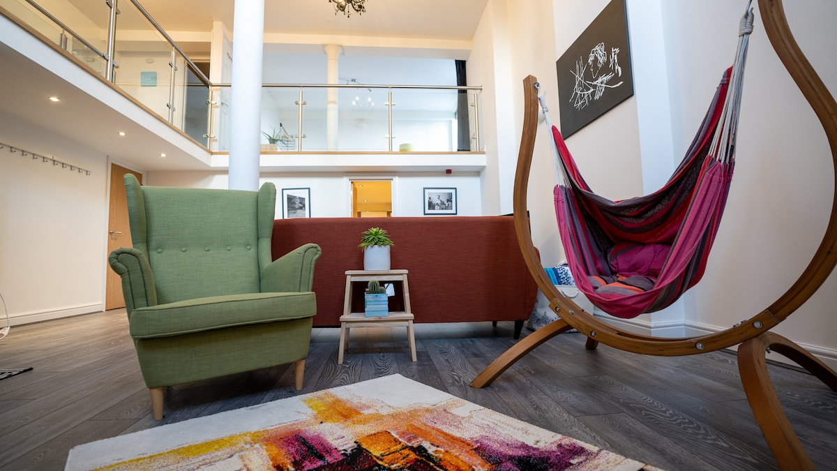 The open-plan living area features double-high ceilings and views of the mezzanine. A green armchair and a stylish hammock are included, along with a brown sofa. A small side table is positioned near a decorative plant, complemented by a vibrant area rug on the wood floor.