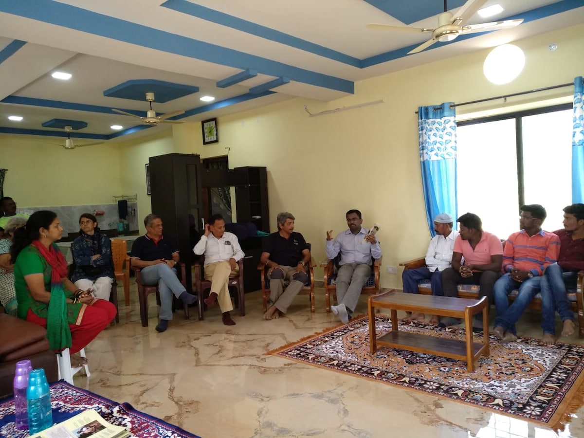 A group of individuals is seated in a spacious room, arranged in a semi-circle. The room features light-colored walls and a large window with blue curtains, letting in natural light. A patterned rug covers the floor, and a coffee table is centrally placed.