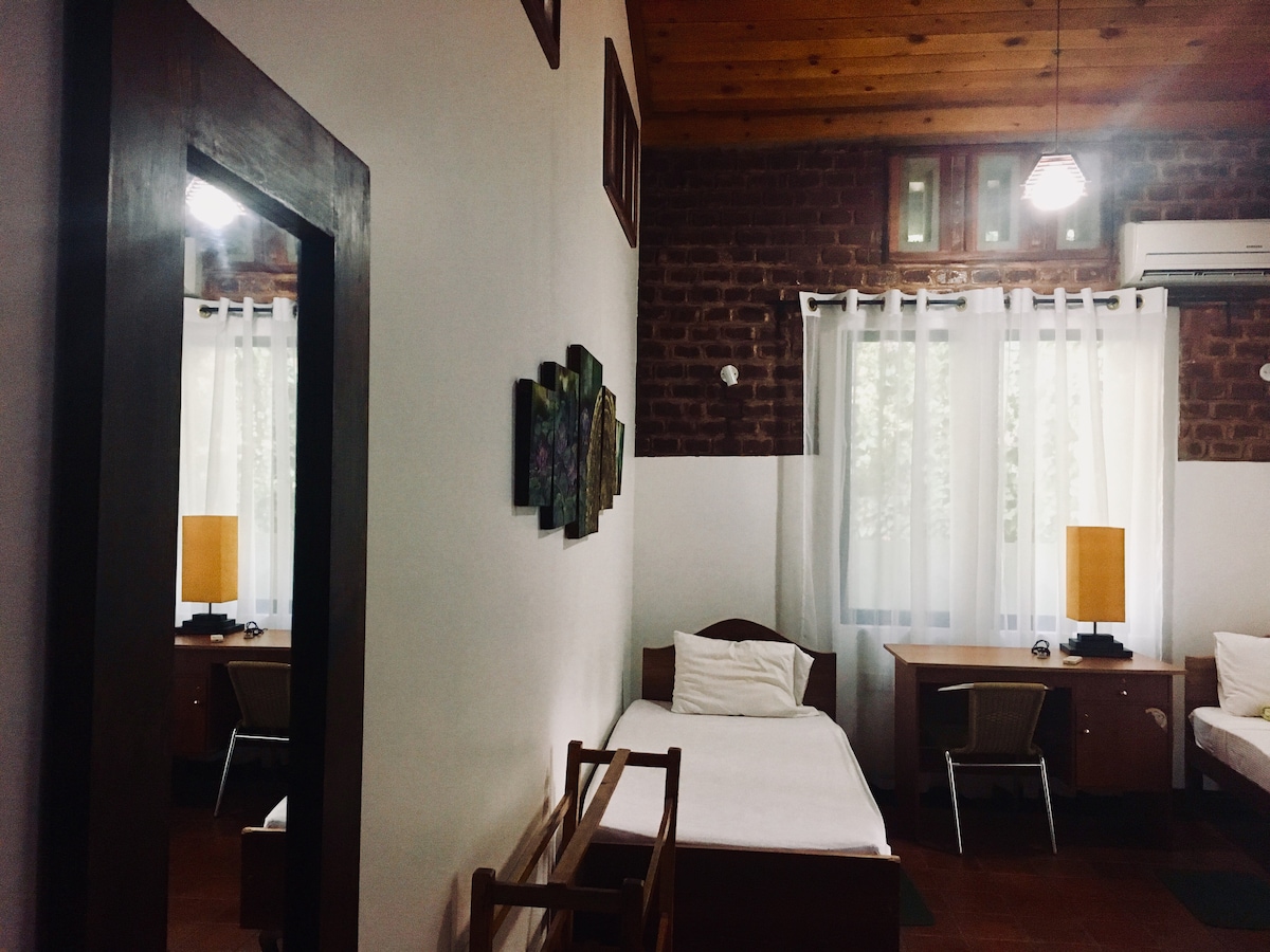 The room features warm wooden beams and brick accent walls, creating a rustic atmosphere. Soft light filters through sheer curtains, highlighting two beds and a desk area. Decorative artwork adorns the wall, while a mirror reflects the inviting space.