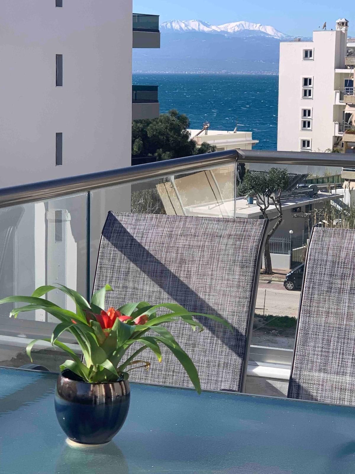 A modern balcony features two grey chairs positioned around a glass table. A potted plant with red blooms adds a touch of color, while the blue sea and distant snow-capped mountains create a scenic backdrop.