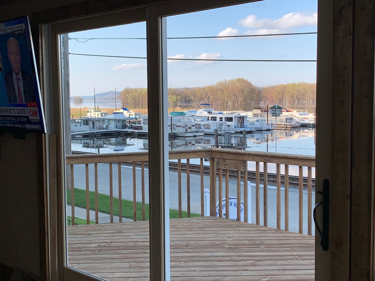 Large glass doors reveal a view of a marina with docked boats and calm waters. The deck, made of natural wood, creates an inviting space for relaxation. Lush greenery is visible in the background, complementing the serene riverside setting.
