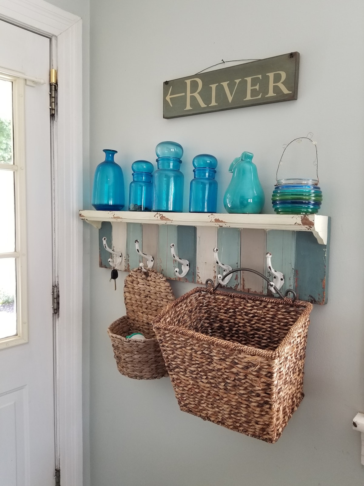 A charming entryway features a wooden shelf adorned with various blue and green glass containers. Below, hooks are provided for hanging items, and woven baskets offer additional storage. Light is filtered through the nearby door, enhancing the welcoming atmosphere.