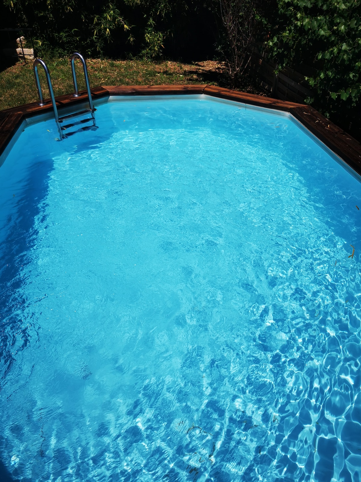 A swimming pool is surrounded by a wooden deck, featuring a ladder leading into the water. The pool is filled with clear blue water, reflecting sunlight and creating gentle ripples on the surface. Lush greenery surrounds the pool area, providing a sense of privacy.
