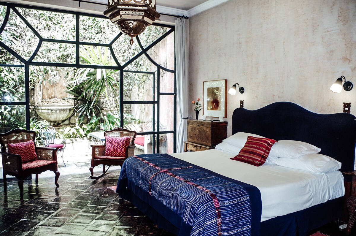 A spacious bedroom features a king bed dressed in crisp linens, with a decorative blanket and an antique writing table. Two antique cane chairs provide seating. Large wrought iron and glass French doors open to a private garden, allowing natural light to fill the room.