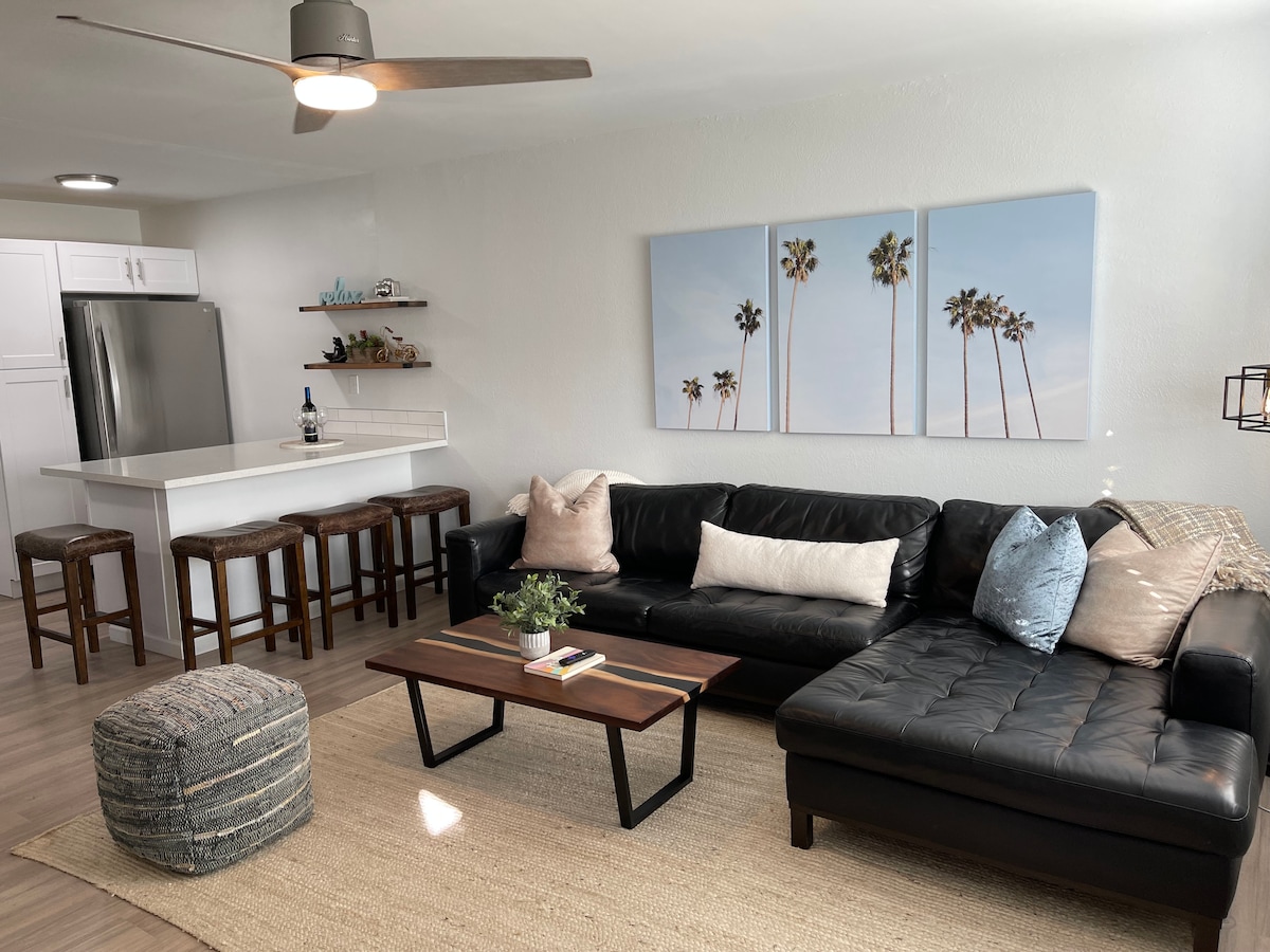 A bright living area features a black leather sectional sofa with decorative pillows. A coffee table made of wood and metal stands in front, accompanied by a pouf. A modern ceiling fan hangs above, while palm tree artwork adds a touch of the outdoors. A dining area with stools completes the space.