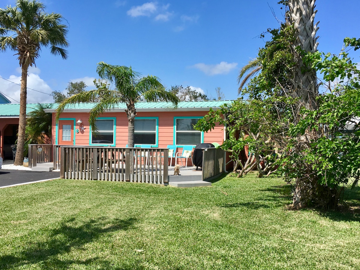 The exterior of the cottage is shown, featuring light coral-colored walls and a metal roof. Green palm trees and shrubs surround the property, with a deck visible in front, furnished with seating. A well-maintained lawn stretches across the scene under a clear blue sky.