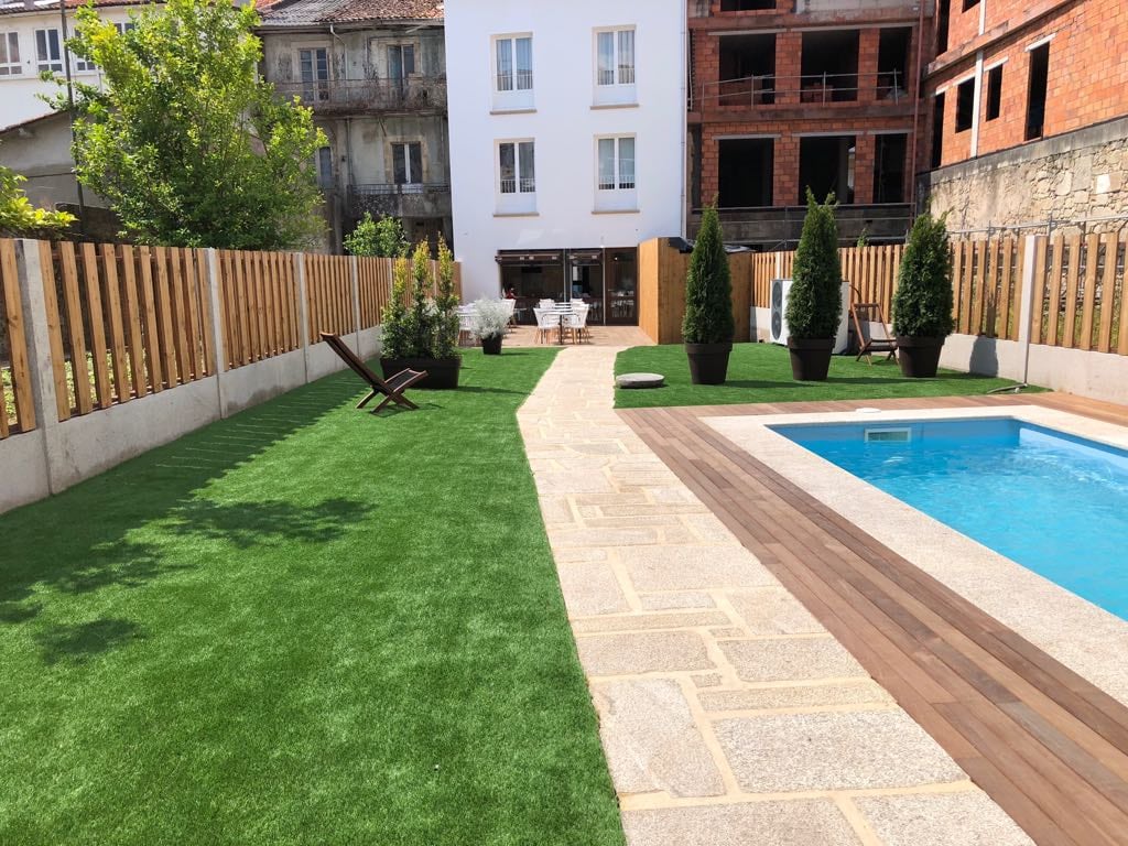 A well-maintained outdoor space features a neatly manicured lawn beside a swimming pool. Surrounding the area are tall shrubs and decorative trees. Comfortable lounge chairs are positioned on the grass, and a paved pathway leads to the entrance of the hotel, enhancing the overall functionality of the space.