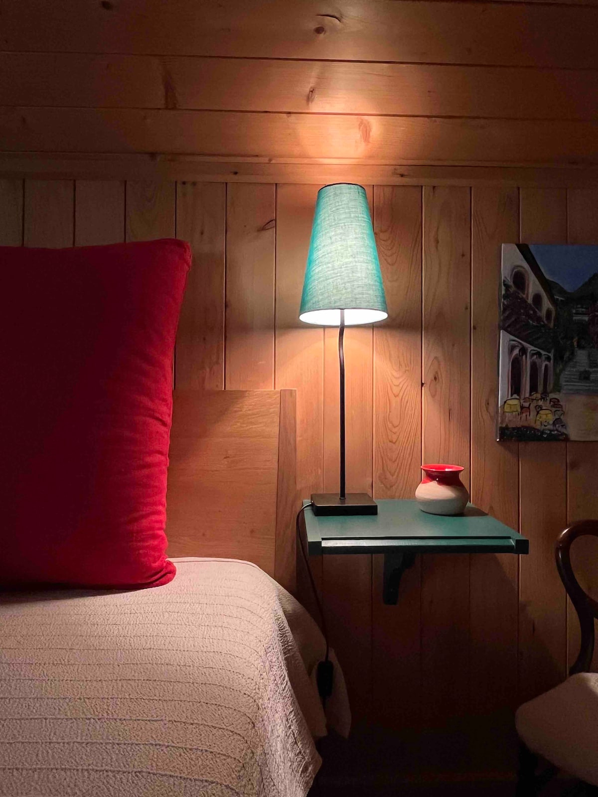 A warm sleeping area features a wooden wall, complemented by a spacious bed with a textured light blanket and a large red pillow. A small bedside table holds a lamp with a turquoise shade and a ceramic vase, next to a colorful wall art piece.