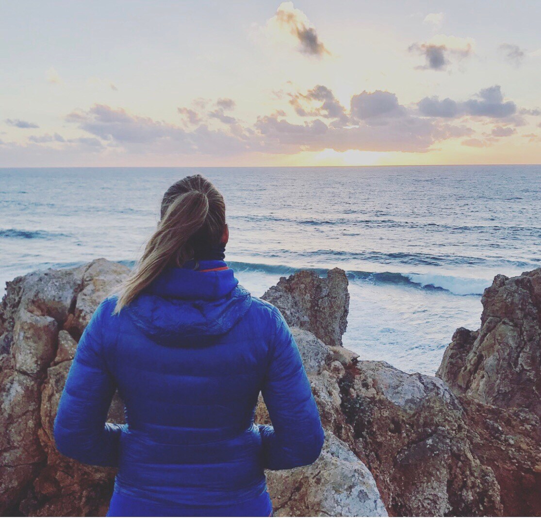 The image captures a person standing on a rocky outcrop overlooking an expansive ocean. Waves gently crash against the rocks under a colorful sunset, with hues of orange and blue blending in the sky. The individual is dressed in a blue jacket, facing the horizon.