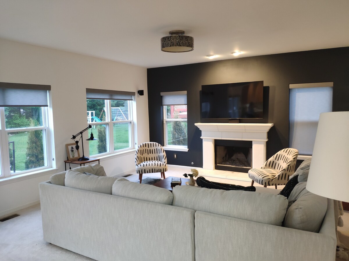 A cozy living room features a light-colored sectional sofa facing a modern fireplace. Two patterned chairs are positioned nearby, and a large flat-screen TV is mounted above the mantel. Natural light enters through multiple windows, illuminating the space.