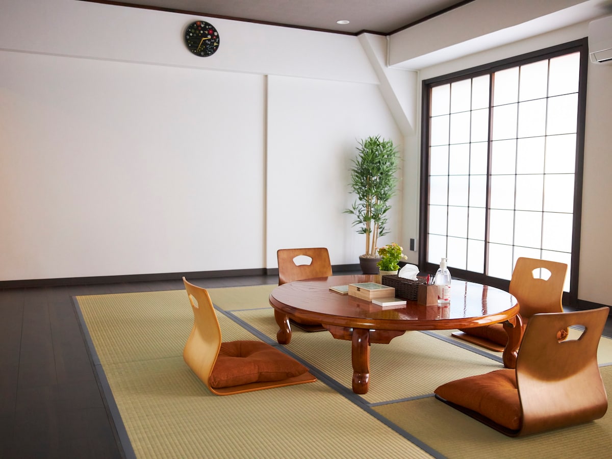 A traditional Japanese-style room features tatami flooring and a round wooden table at its center. Four floor cushions are arranged around the table. Natural light filters through large sliding doors, while a decorative plant adds a touch of greenery to the space.