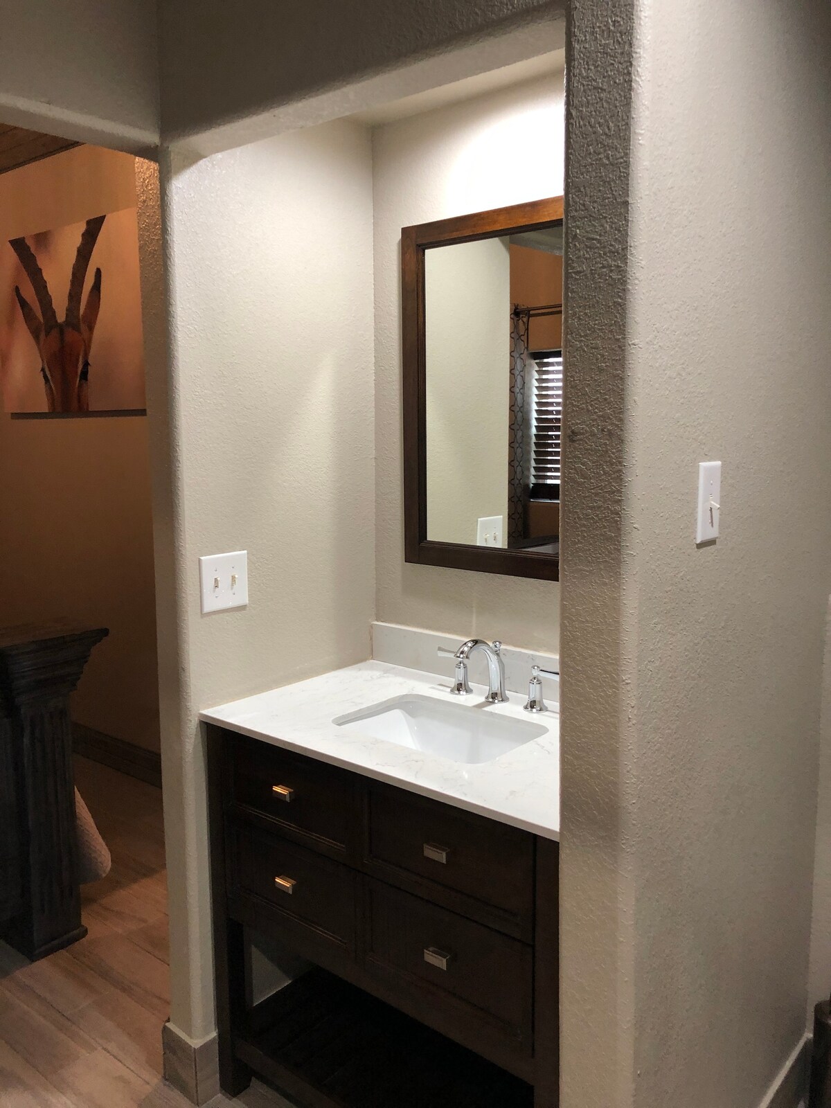 A compact bathroom space is visible, featuring a modern sink with chrome faucets, set atop a light-colored countertop. Below the sink, a dark wood cabinet provides storage with two drawers. A rectangular mirror hangs above the sink, reflecting the neutral walls.