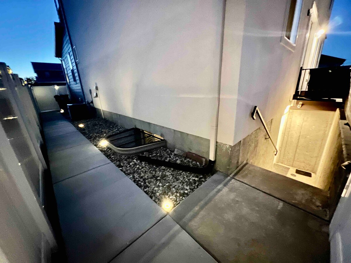 A well-lit pathway is shown alongside a home's exterior, featuring smooth concrete walkways and bordered by decorative gravel. Soft ground lighting illuminates the path, providing a welcoming ambiance. Stairs lead down toward a lower entry, ensuring easy access to the basement apartment.