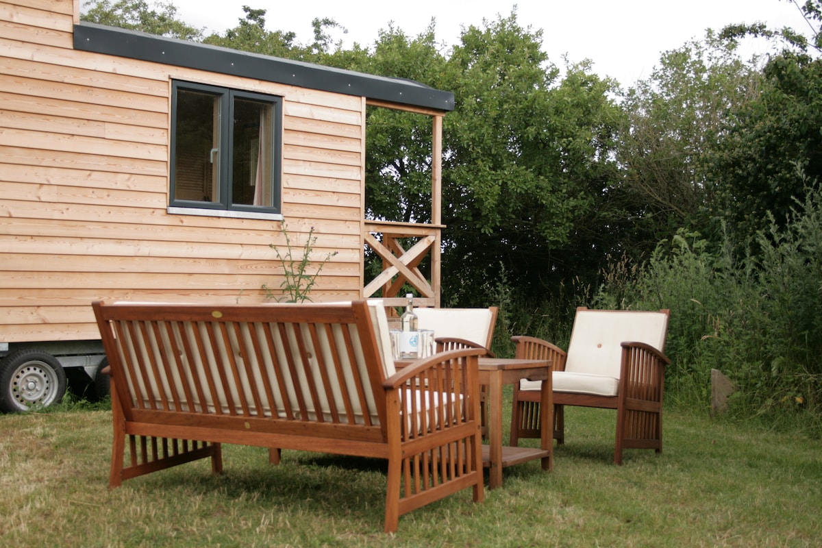 An outdoor seating area features a wooden sofa set consisting of two armchairs and a sofa, positioned around a small coffee table. The structure stands amidst lush greenery, with a wooden tiny house visible in the background.