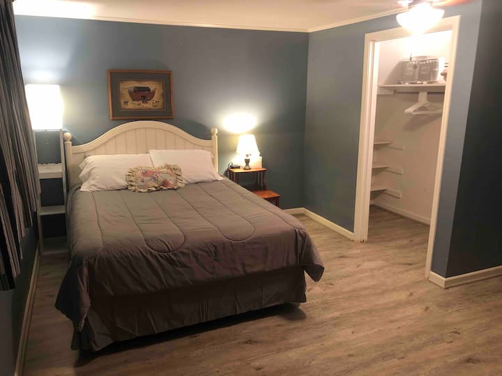 Bedroom with queen bed, charging stations and walk-in closet with extra blanket, hangers and a fan.