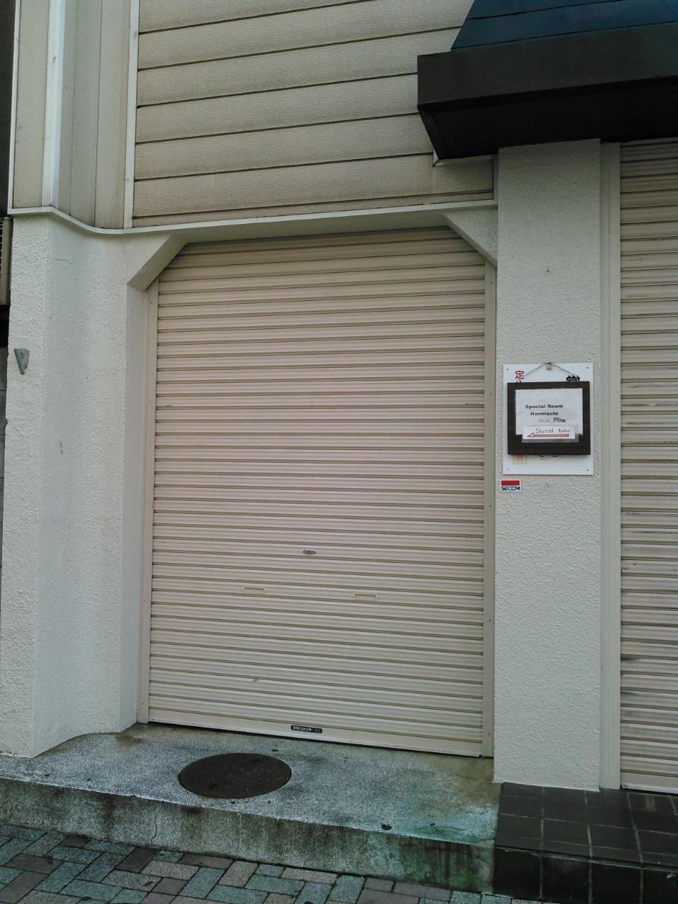 An exterior view of a closed roll-up door is visible. The door is beige with a textured surface. A small sign is placed beside the door, providing information about the space. The surrounding wall is a light color, complementing the door.