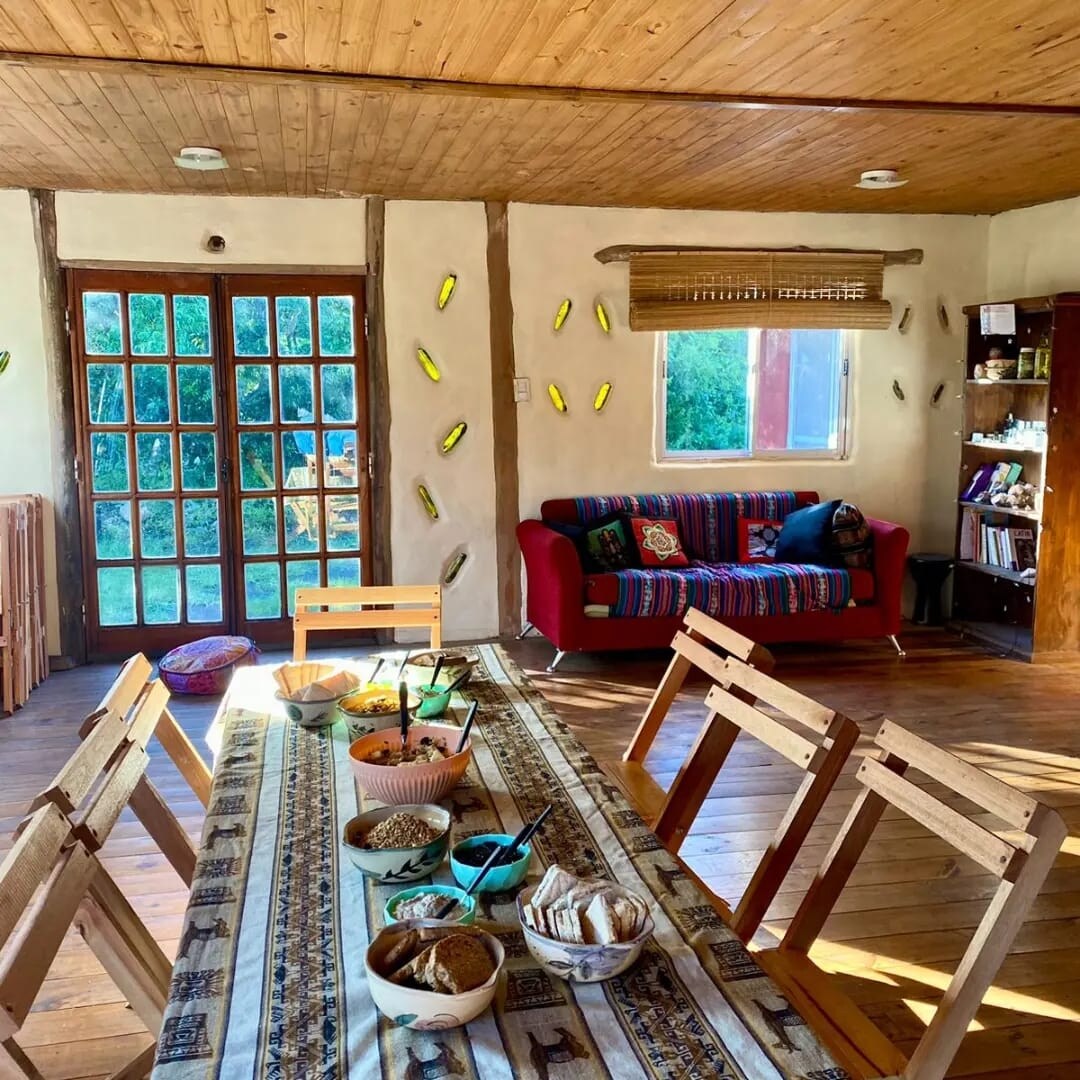 A spacious living and dining area is presented, featuring a long wooden table surrounded by folding chairs. A cozy sofa with colorful cushions sits nearby. Natural light floods the room through large windows, enhancing the warm wooden tones and inviting atmosphere.