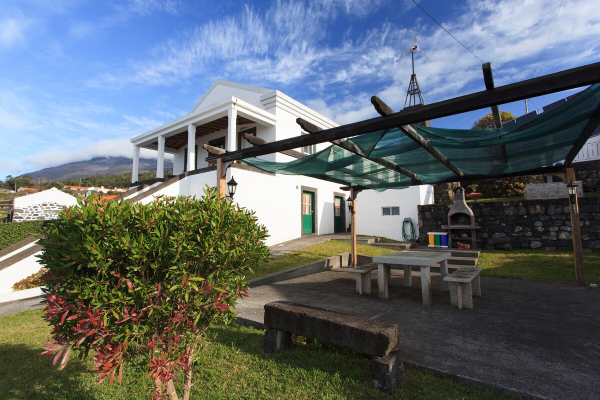 An outdoor patio area features a large stone table surrounded by benches, shaded by a green canopy. In the background, a white building with multiple columns stands against a blue sky. A hint of mountains is visible in the distance, complementing the serene landscape.