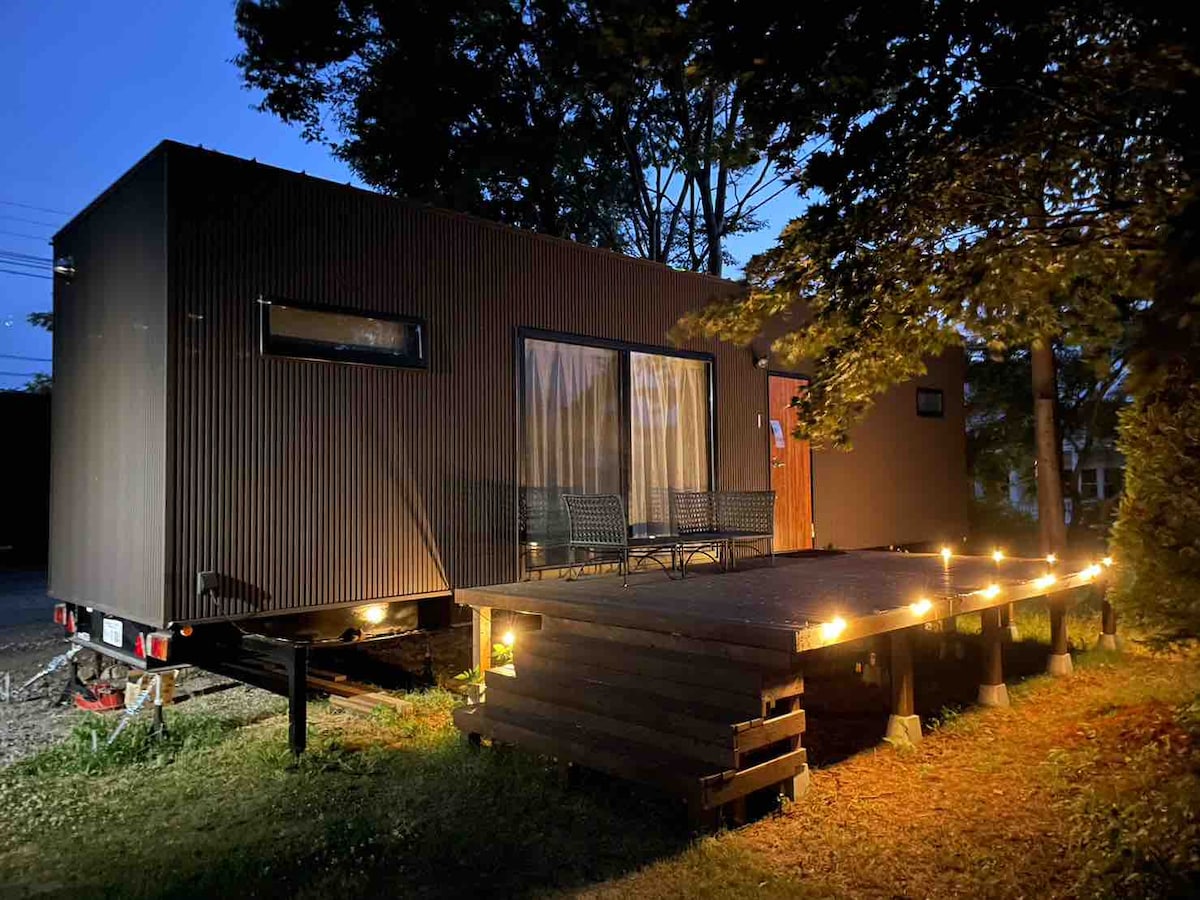 An evening view of a modern tiny house is presented, featuring sleek black exterior walls and large windows. A wooden deck is illuminated by soft outdoor lights, and comfortable chairs are positioned on the deck, surrounded by natural greenery.