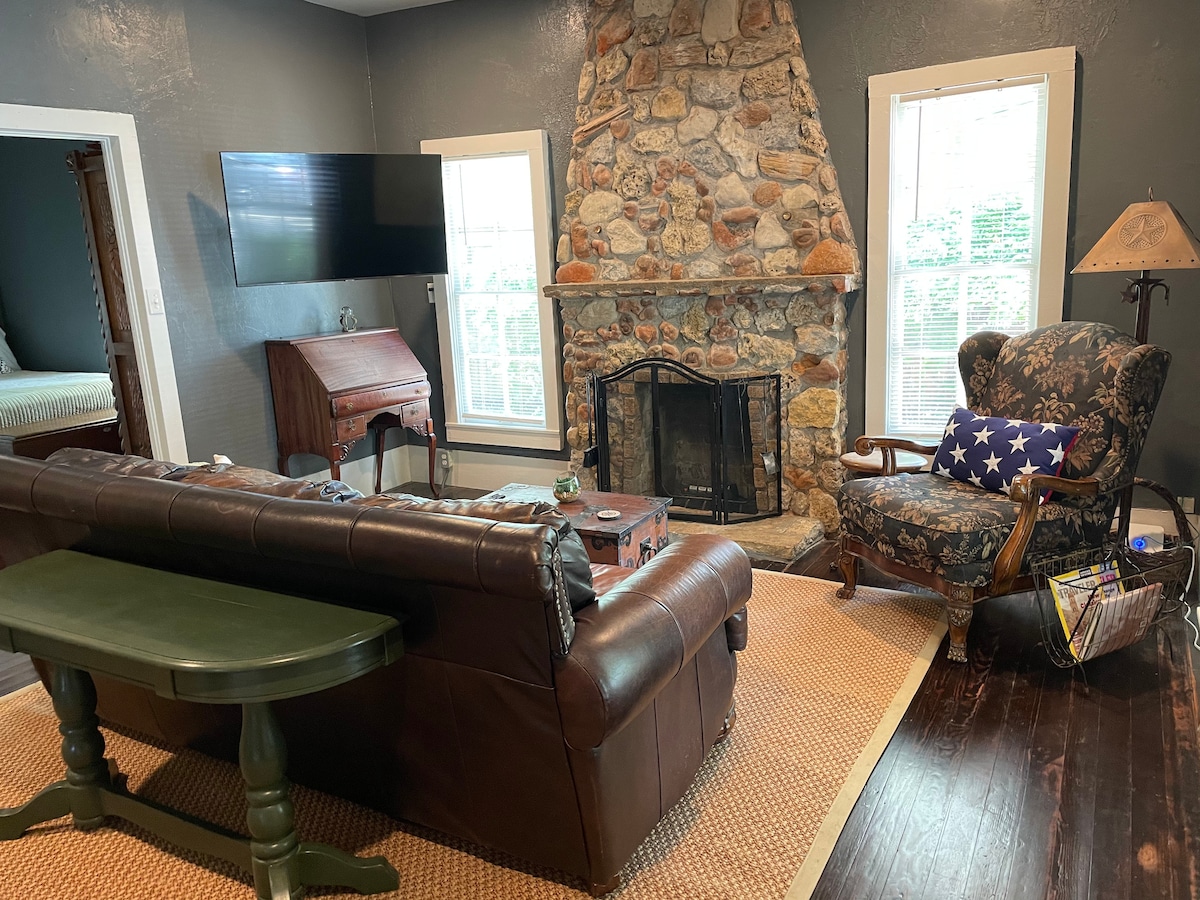 A living area is highlighted by a stone fireplace and a large flat-screen television mounted on the wall. A comfortable leather sofa and an armchair with a patterned fabric provide seating. Natural light enters through the windows, illuminating the rustic decor.