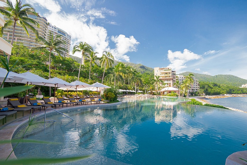 A large swimming pool surrounded by lounge chairs is seen, with tall palm trees lining the area. The water reflects the clear blue sky and buildings in the background. Sun umbrellas provide shade over the seating, enhancing the serene atmosphere of the tropical resort.