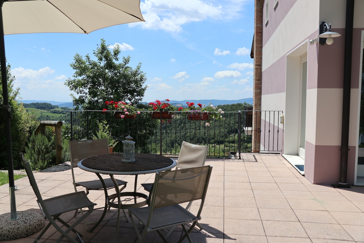 A spacious terrace area is shown, featuring a round table surrounded by four chairs. A parasol provides shade, while vibrant flower boxes adorn the railing. The backdrop showcases a panoramic view of rolling hills under a clear blue sky.