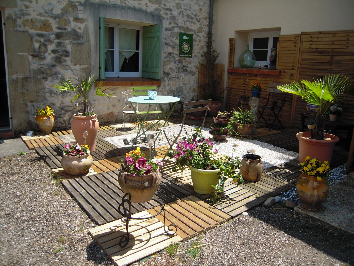 A charming outdoor patio features wooden decking surrounded by an array of potted plants and flowers. A small round table with chairs is positioned near the entrance, while larger pots add greenery to the space. The atmosphere is inviting and well-maintained.