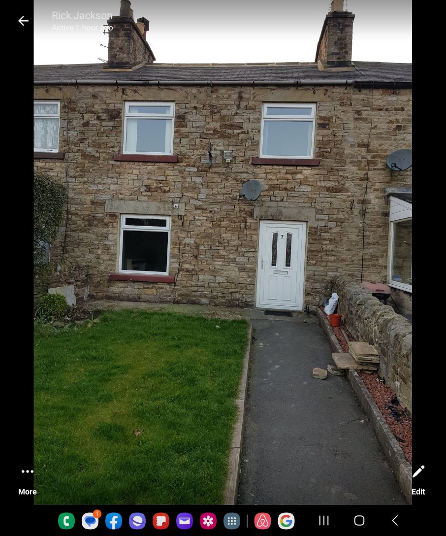 The exterior of a stone cottage is presented, featuring a neatly maintained front garden with a patch of grass and a stone pathway. The entrance door is centered, flanked by two windows. Satellite dishes are visible on either side of the upper windows.