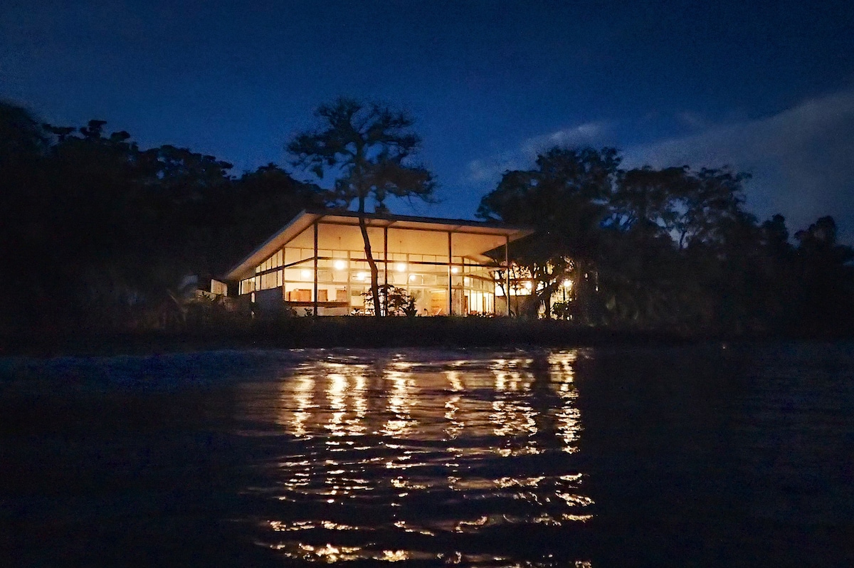 The modern architectural home is viewed from the water at night, illuminated by warm lights from within. Reflections shimmer across the calm surface of the water, while the surrounding trees are softly silhouetted against the night sky.