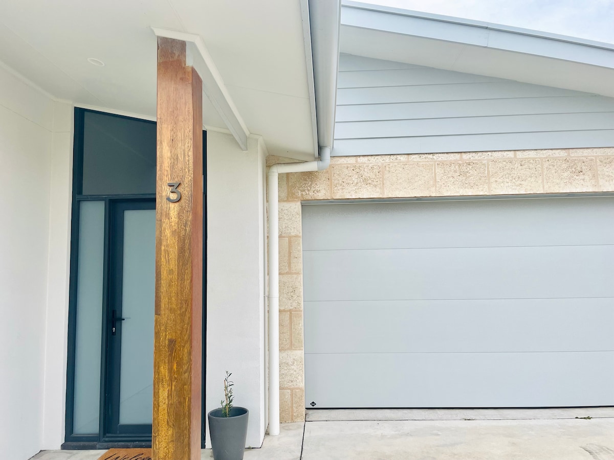 The entrance of the house features a wooden column beside a contemporary front door, inviting guests inside. A decorative potted plant adds a touch of greenery, while the garage door presents a sleek design, complementing the light-colored exterior.