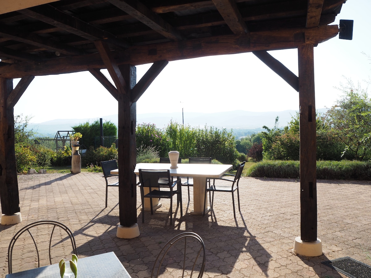 A spacious terrace is featured, supported by sturdy wooden beams. A large table is accompanied by several chairs, providing an inviting space for relaxation. Lush greenery and distant hills create a scenic backdrop, enhancing the outdoor experience.