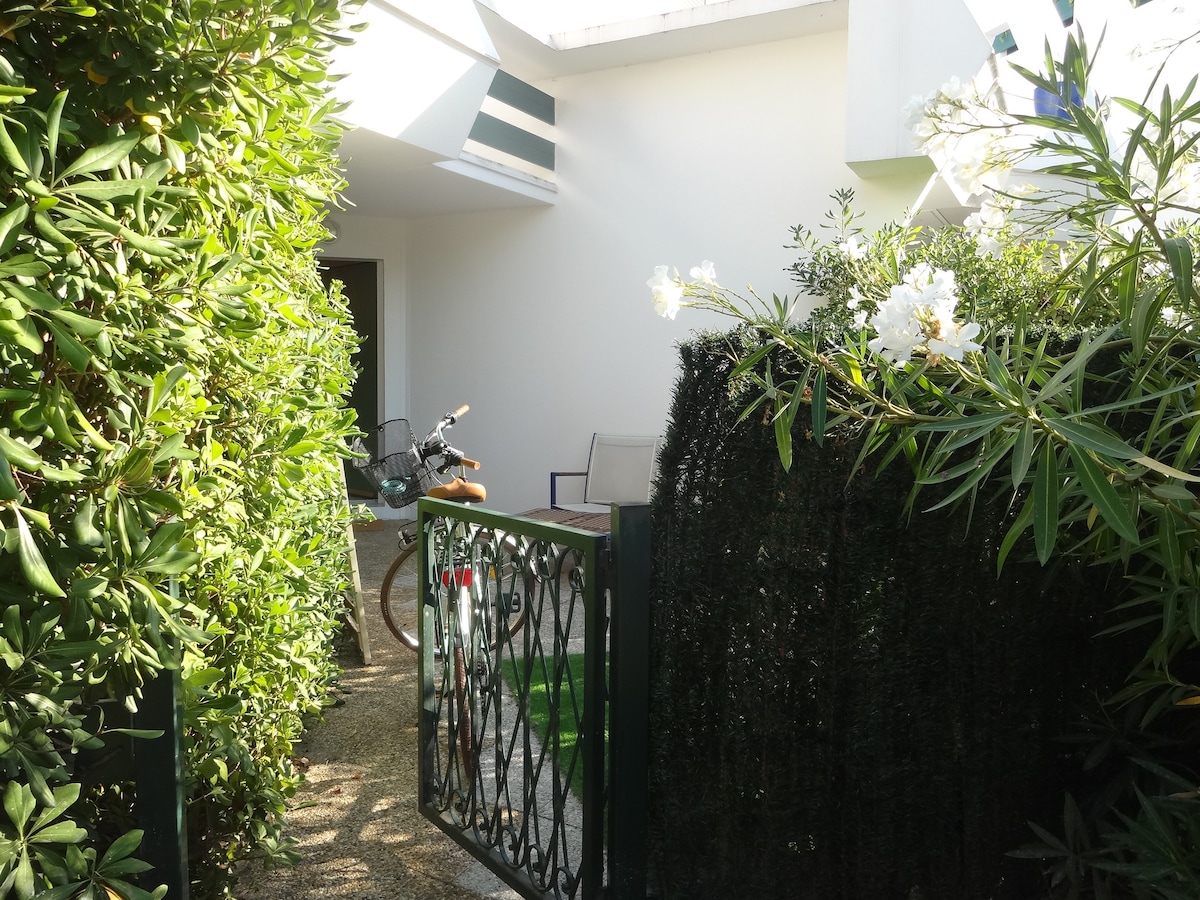The entrance is framed by lush greenery, leading to a private outdoor area. A bicycle is parked beside a modern chair, indicating a welcoming space. A gate is visible, providing access to the property, and soft sunlight illuminates the pathway.