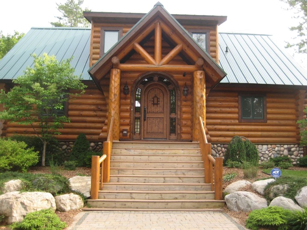 The entrance of a custom log cabin is presented with large, natural wood steps leading up to a double door framed by stone accents. A peaked roof with overhanging eaves and wooden columns emphasizes the rustic design. Surrounding greenery and decorative boulders enhance the natural setting.