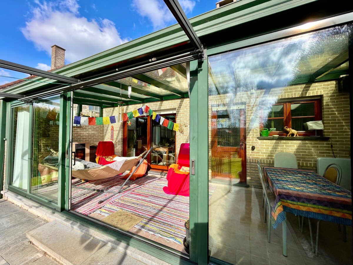 A spacious veranda is surrounded by large glass panels, offering ample natural light. The area features a colorful woven rug, a dining table, and two comfortable chairs. Decorative fabric banners hang across the space, adding a touch of vibrancy to the inviting setting.