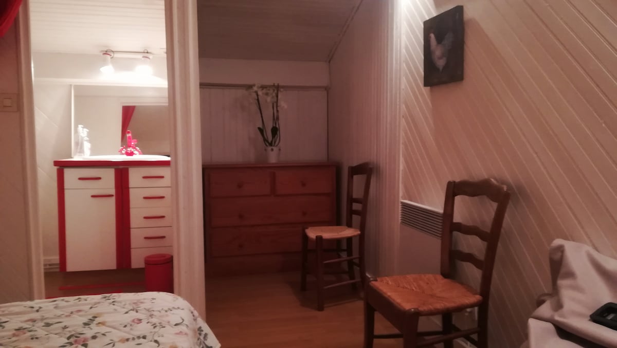 An interior view showcases a private room with a decorative wall and wooden flooring. Two wooden chairs are positioned near a dresser, which features a smooth surface. A small vanity area with lighting is visible in the background, along with a decorative wall piece.