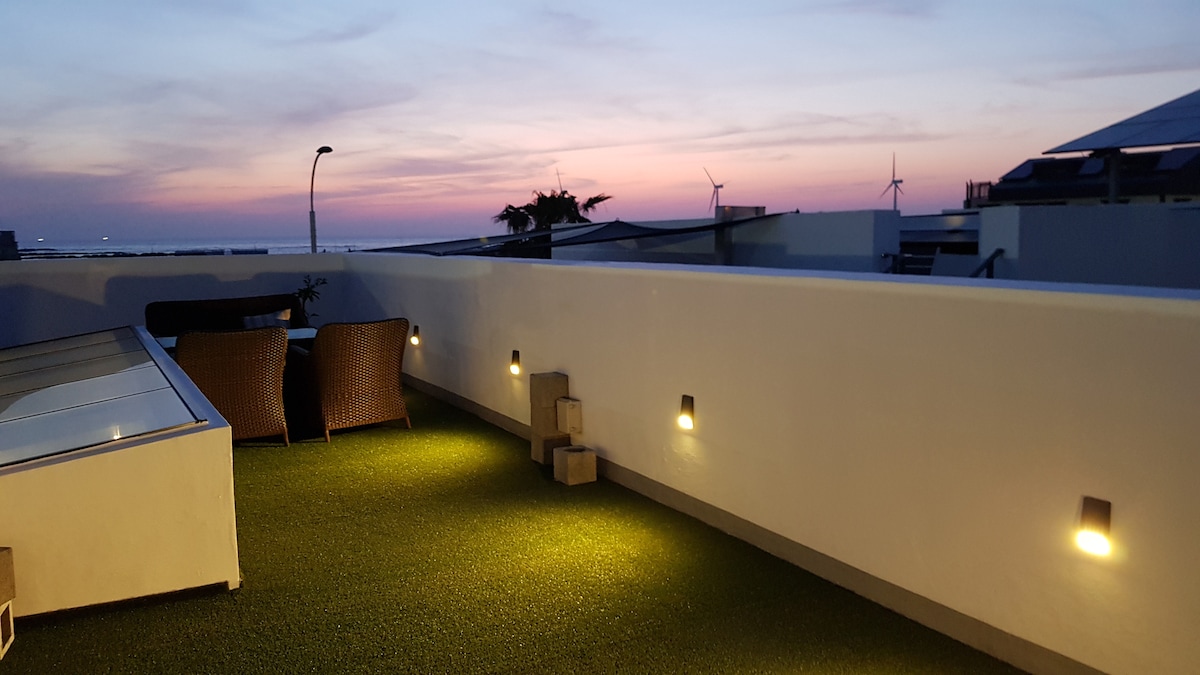 A spacious rooftop area is illuminated by soft outdoor lighting, highlighting a serene view of a sunset with pastel hues in the sky. Comfortable seating arrangements are provided, overlooking distant wind turbines and the surrounding landscape.