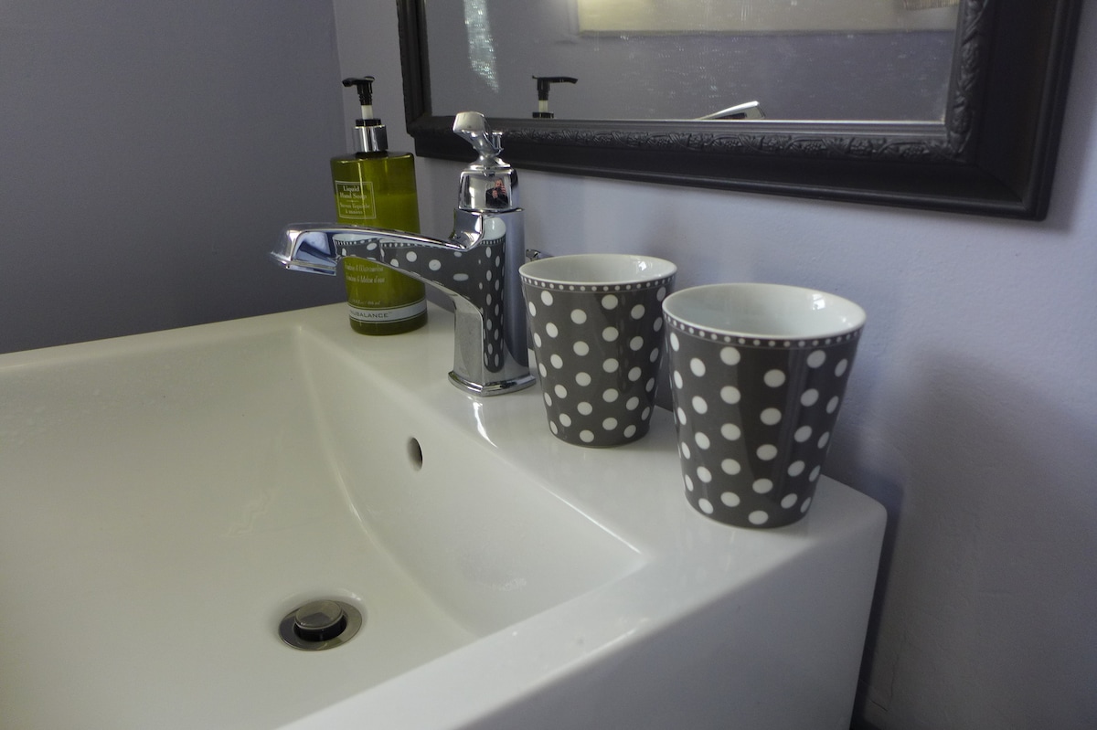 A modern bathroom sink area is shown with a sleek faucet and a polished countertop. Two polka-dotted cups are placed beside a pump bottle of green liquid soap. The reflection of a framed mirror can be seen in the background, enhancing the space's brightness.