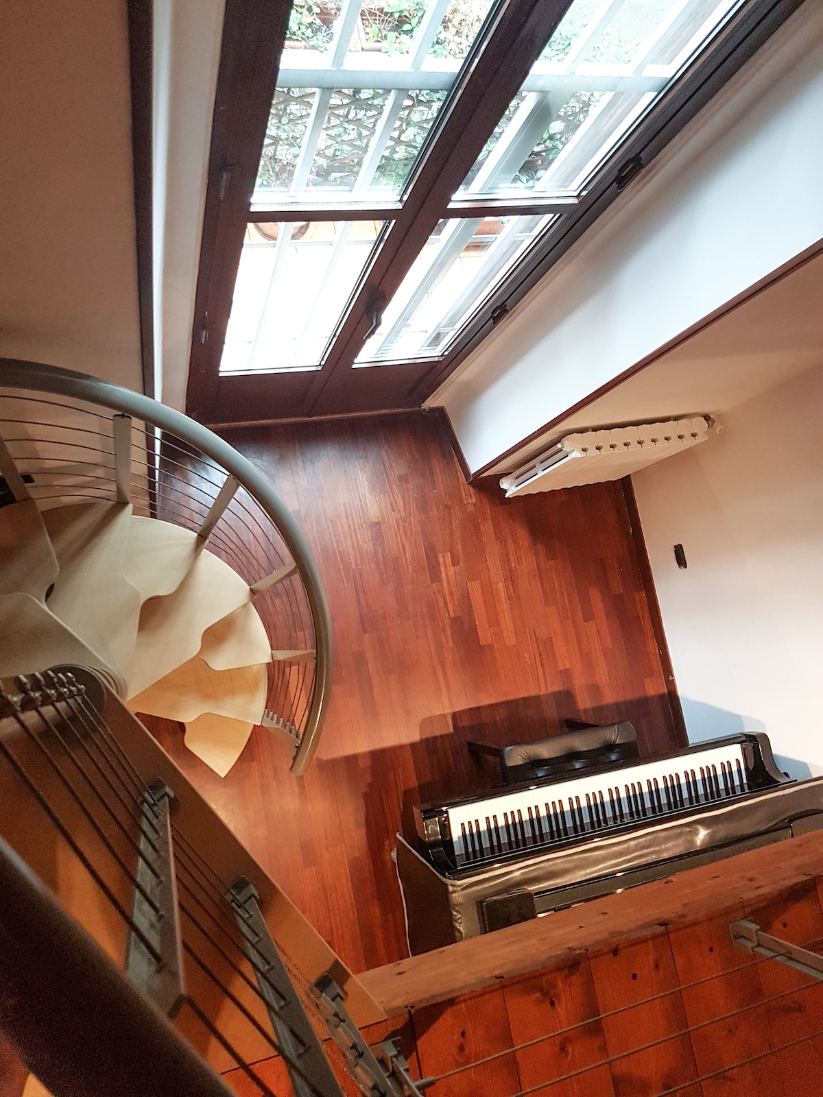 A spiral staircase leads to a cozy area featuring a piano on polished wooden flooring. Large windows allow natural light to fill the space, while a view of greenery is visible through the glass panes. The overall ambiance is serene and inviting.