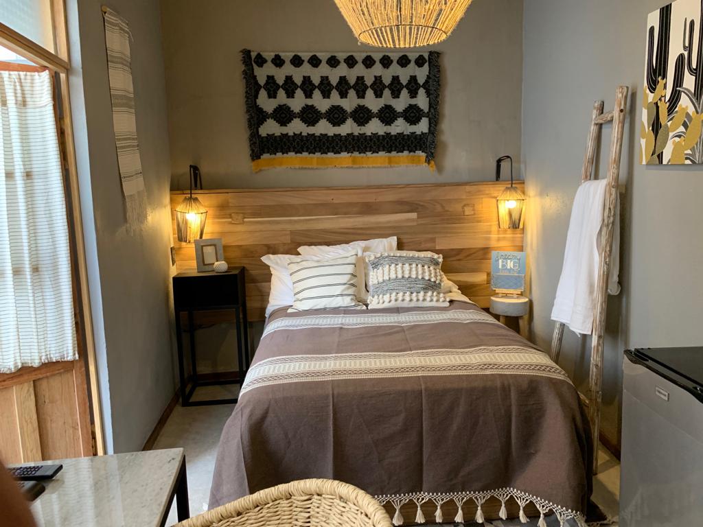 A cozy sleeping area is featured with a rustic wooden headboard and warm lighting from two wall sconces. A spacious bed is dressed in neutral tones, complemented by decorative cushions. Natural textures are present in the woven wall art and nearby furniture.