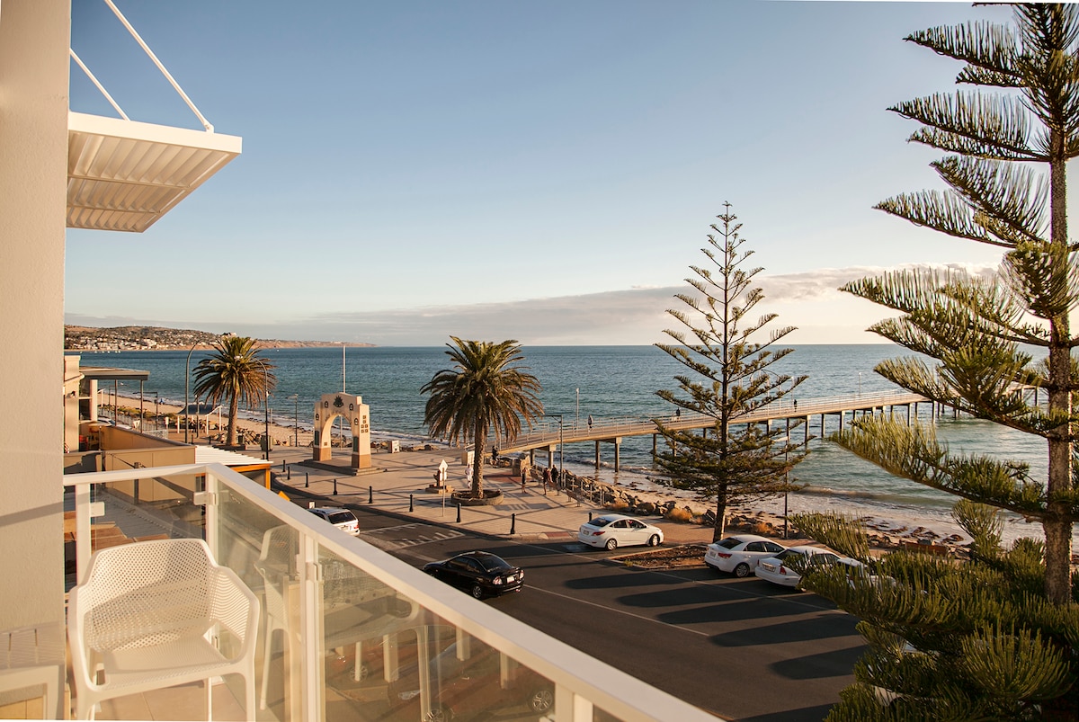 A view from a balcony showcases a vibrant coastline along Brighton beach. Soft waves gently lap at the shore, while palm trees line the promenade. The horizon reveals a calm sea under a pale sky, and local walkways are visible with pedestrians enjoying the scenery.